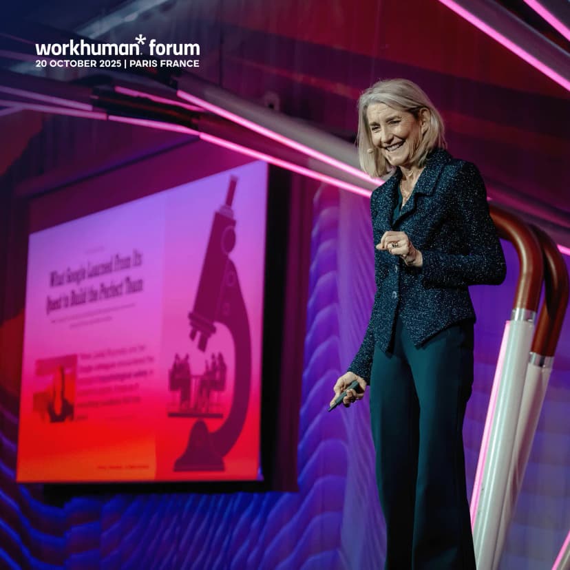 A woman with light hair is seen speaking on stage at the Workhuman Forum event in Paris on October 20, 2025. She is wearing a sparkly dark jacket and holding a pen, smiling while engaging with the audience. Behind her, a projection displays text that begins with "What Google Learned From Its Effort to Build Its Paycheck," alongside an image of a microscope and people in discussion. The stage is adorned with colorful lighting that adds to the vibrant atmosphere of the event.