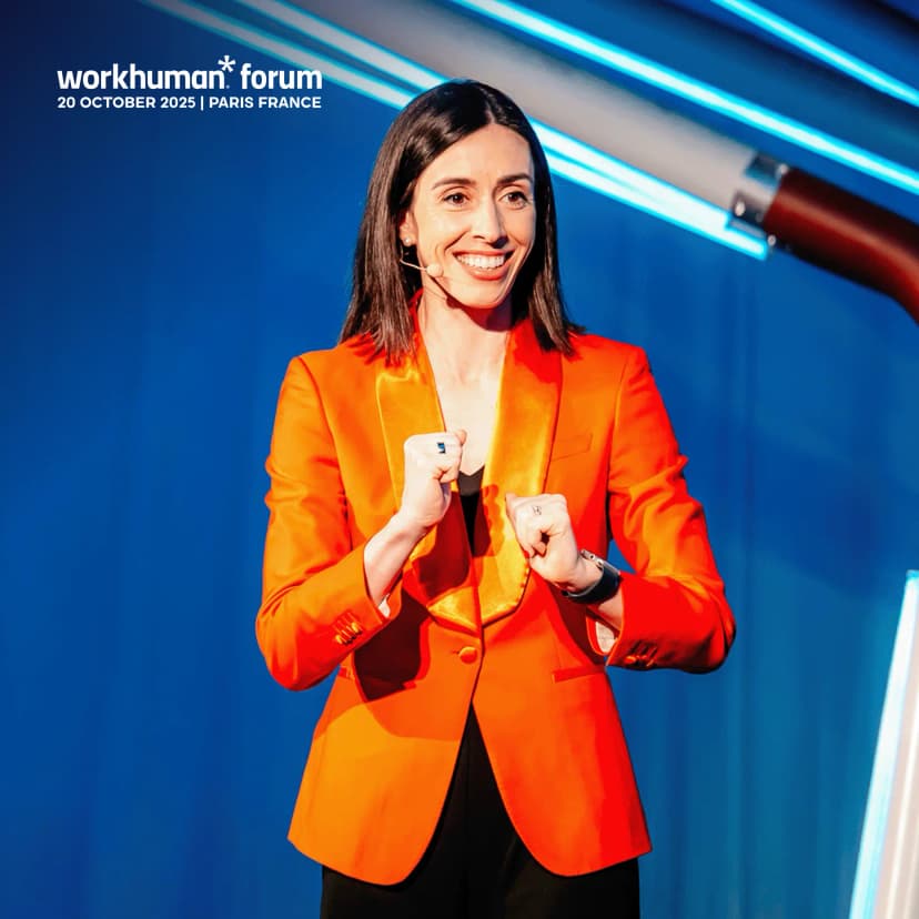 A woman in a vibrant orange blazer stands on stage, smiling and gesturing with her hands in a positive and engaging manner. She has long dark hair and is positioned in front of a blue backdrop with bright lighting accents. The text overlay indicates the event as the "workhuman forum" happening on "20 OCTOBER 2025 | PARIS FRANCE."