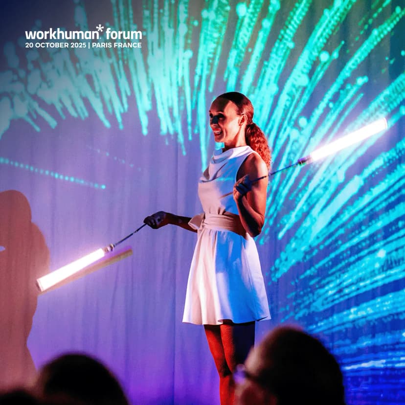 A woman in a white dress with a belt is performing with lighted props on stage at the Workhuman Forum event. She is smiling, and colorful light streaks are projected in the background, creating a vibrant atmosphere. The image includes text at the top indicating the event title and date: "workhuman* forum 20 OCTOBER 2025 | PARIS FRANCE."