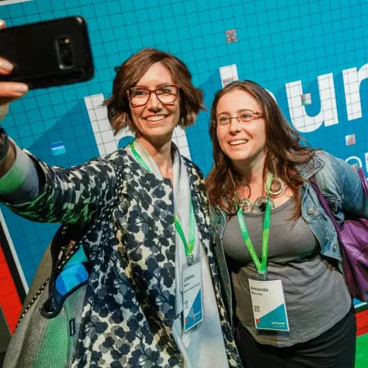 Two women are smiling and taking a selfie in front of a colorful, patterned backdrop that features the word "Human" prominently. They are wearing name tags and have green lanyards around their necks. Both women are dressed casually, with one wearing a patterned coat and the other a gray top. The scene captures a moment of joy and connection at an event.