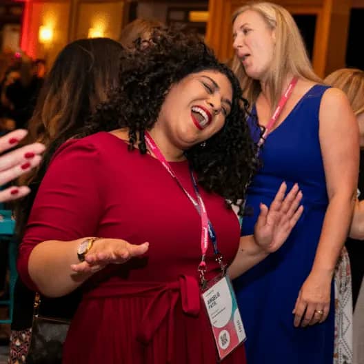 A woman with curly hair is joyfully dancing and smiling while wearing a red dress. She has a name tag visible around her neck. In the background, a woman wearing a blue dress is seen engaged in conversation with another group. The setting appears to be festive, suggesting a lively event.