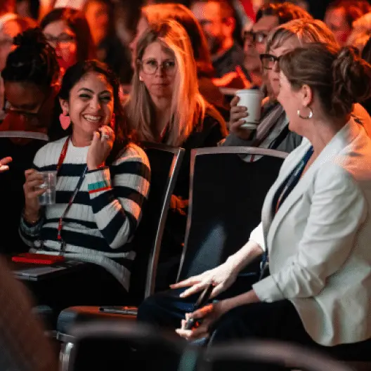 A diverse group of attendees at a conference sits in an audience, engaged in conversation and smiling at one another. In the foreground, a woman wearing striped clothing is laughing and holding a drink, while a woman in a white blazer is gesturing as she speaks. Behind them, other attendees are seen, some holding cups, indicating a lively and interactive environment. The setting appears professionally lit, creating a warm and inviting atmosphere.