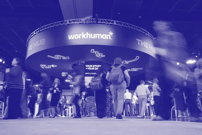 A busy event space with attendees walking around under a large circular display for Workhuman, featuring the words "THE HUB" prominently. The atmosphere is bustling, with people engaged in conversation as they navigate the area, which is well-lit and decorated for the occasion. The backdrop also has abstract shapes and arrows, creating an energetic vibe.