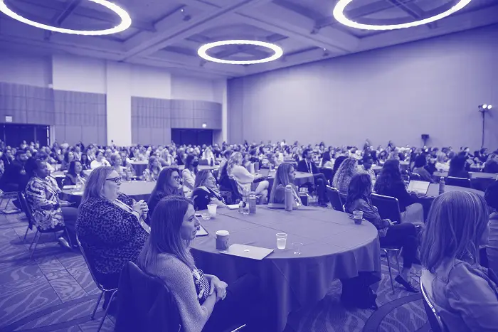 A large audience of attendees sits at round tables in a conference setting, listening attentively. The room is decorated with modern lighting fixtures, and many participants are engaged with their devices or taking notes. There are cups and materials scattered on the tables. The atmosphere appears focused and active, indicative of a collaborative and educational event.