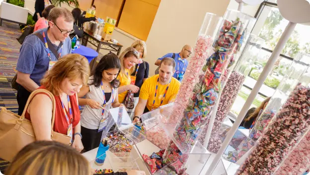 A group of attendees at a conference are gathered around a candy display featuring various colorful treats in clear containers. People are engaging with the candy, selecting items while wearing lanyards. The background shows a bright setting with tables and other activities, creating a lively atmosphere.