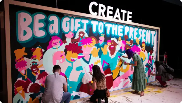 A colorful mural being created at an event, featuring the phrase 'BE A GIFT TO THE PRESENT' in bold lettering at the top. Several artists, including individuals wearing various outfits, are engaged in painting vibrant characters and scenes on the wall. The atmosphere appears lively and creative, with a focus on collaboration and artistic expression.