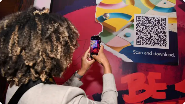 A person with curly hair is holding a smartphone up to a colorful wall graphic that features a QR code. The graphic also contains the text 'Scan and download.' The wall design incorporates various shapes and colors, enhancing the overall visual appeal.
