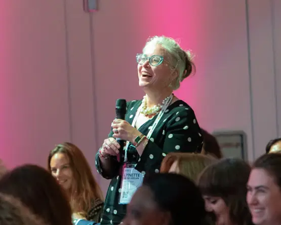 A group of three women poses in front of a colorful backdrop that reads "THE FUTURE OF WORK IS HUMAN." The women are smiling and dressed in professional attire, and the vibrant wall features shades of teal and pink with a diamond shape. The setting appears to be a conference or event.