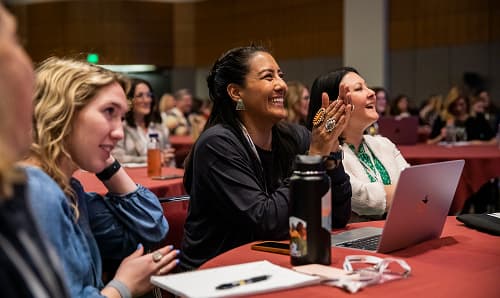A group of engaged attendees at a conference, including a woman with long dark hair smiling and clapping, while others around her laugh and show interest. Several laptops and water bottles are visible on the tables in front of them, creating a vibrant atmosphere of participation and enthusiasm.