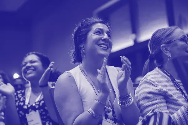 A group of people attending an event, with one woman in the foreground smiling and applauding. The image has a blue filter applied, creating a lively and engaging atmosphere. Other attendees can be seen in the background, also enjoying the moment. The overall scene conveys a sense of joy and participation at a shared experience.