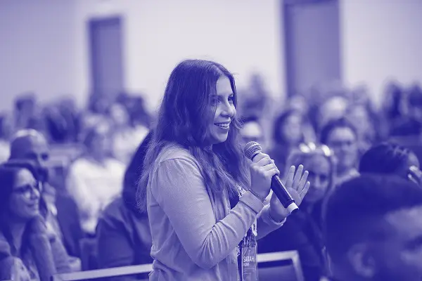A woman with long hair is smiling while holding a microphone, engaging with the audience in a conference setting. In the background, several attendees are visible, seated and facing her, indicating an interactive discussion or Q&A session. The image has a blue overlay, giving it a vibrant, dynamic feel.