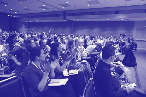 A large audience is seated in a conference room, actively applauding with smiles and engagement. Various attendees can be seen, some holding papers or notepads in their laps, while others are focused on the front of the room, indicating a lively atmosphere of interaction and appreciation during the event. The setting suggests a professional gathering, likely involving presentations or discussions.