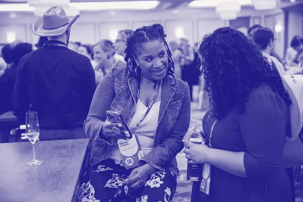 A woman in a denim jacket smiles while holding a smartphone, engaging in conversation with another woman dressed in a dark top. They are among a crowd at a social event, with a glass of champagne on the table next to them. The setting appears to be a well-lit indoor venue.