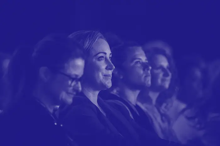 A diverse group of audience members is seated, captivated by a presentation. A woman in the foreground, dressed in a blazer, has a thoughtful smile, while others around her express varying emotions. The image is presented with a blue tint.