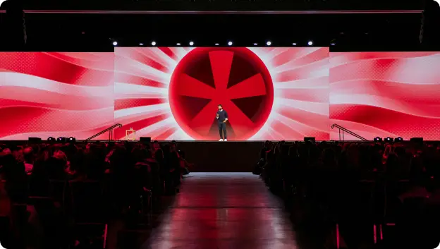 A speaker stands on stage in front of a large backdrop featuring a vibrant red design with radiating lines and a circular symbol. The audience is visible in the foreground, seated and engaged with the presentation.