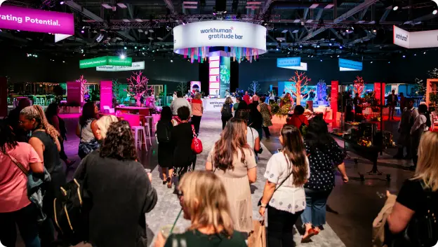 A group of attendees walking towards the 'Workhuman Gratitude Bar' at the conference, surrounded by colorful displays and decorations. The setting features vibrant lighting and decorations that emphasize a celebratory atmosphere. Signs in the background promote themes related to gratitude and potential.