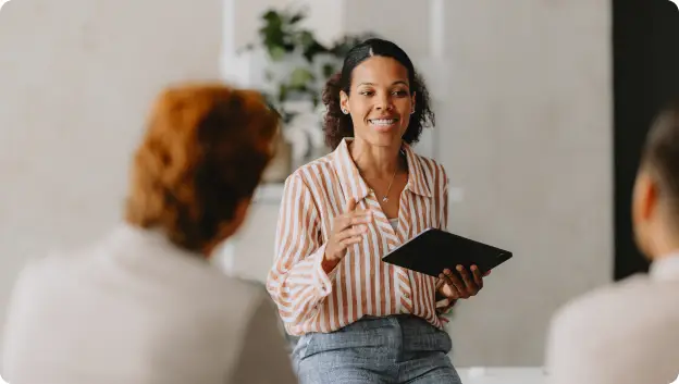 A smiling woman in a striped shirt is engaged in a discussion while holding a tablet. She appears to be addressing two seated individuals who are partially visible, indicating an active conversation or presentation. The setting is bright and professional, with greenery in the background.