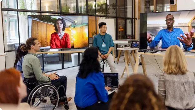 A group of people is participating in a meeting, with some attendees physically present and others appearing on screens. In the foreground, a woman in a wheelchair and another woman wearing a blue shirt are seated, while a man in a green shirt stands in the center. On the screens, individuals are engaged in discussion, one wearing a red outfit and the other in a blue shirt, indicating a virtual collaboration. The space is modern and bright, reflecting a professional setting.