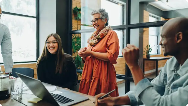 A group of four colleagues in a bright, modern office setting engaged in a lively discussion. Two women, one with long dark hair and glasses, and another with short grey hair and a colorful scarf, are smiling and interacting. A man with short hair is seen from the side, while another man is visible in profile, taking notes. A laptop and some papers are on the wooden table, enhancing the collaborative atmosphere.