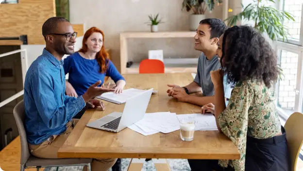 A group of four diverse professionals engaged in a lively discussion around a wooden table. A laptop sits open in front of them, along with scattered papers. The individuals are smiling and appear to be sharing ideas in a modern workspace with plants and natural light in the background.