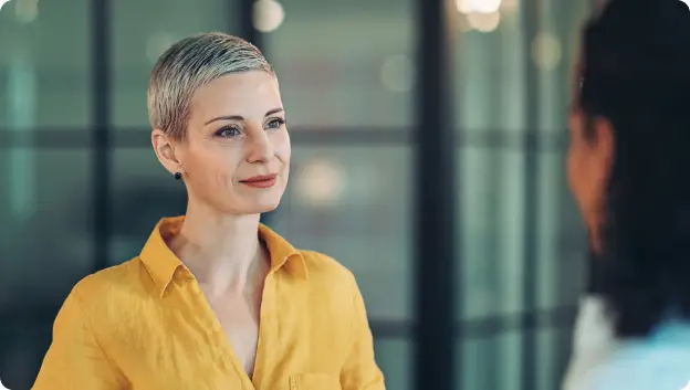A woman with short, light-colored hair wearing a bright yellow shirt listens attentively to another person off-camera in a well-lit indoor setting with blurred background elements.
