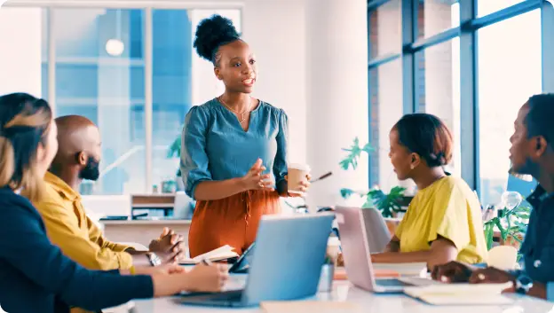 A group of five people are seated around a table in a bright, modern office setting. A woman with curly hair is standing and speaking, holding a coffee cup, while the others listen attentively. They appear engaged in a discussion, and there are laptops and notebooks visible on the table, suggesting a collaborative meeting atmosphere.