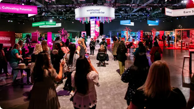 A lively scene at the Workhuman event featuring a 'gratitude bar' display, with attendees interacting and enjoying the atmosphere. The space is decorated with colorful elements and uplifting signs, including phrases such as 'Cultivating Potential' and 'Earn' visible in the background. The crowd consists of people engaged in conversation and activities, creating a vibrant and communal environment.