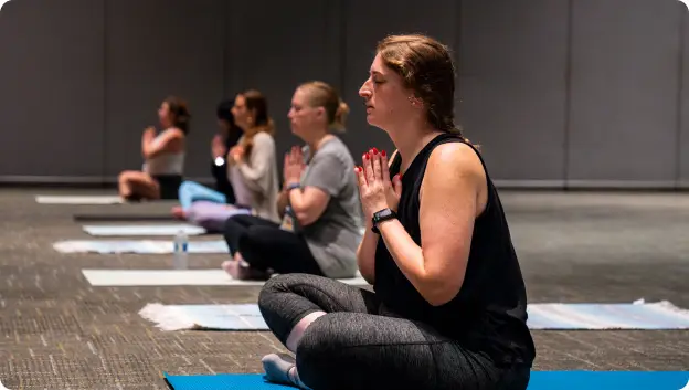 A group of women participating in a yoga session, each seated on mats in a meditative pose with hands in prayer position. They are in a spacious indoor setting with neutral walls and floor. The focus is on one woman in the foreground, who has her eyes closed and appears relaxed, while others are also engaged in meditation.