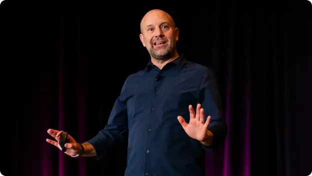 A person stands on stage, gesturing with both hands while speaking to an audience. They are wearing a dark blue shirt and appear to be engaged in a presentation, with a dark backdrop that has hints of purple lighting.