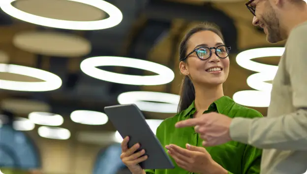 A woman in a green blouse and glasses smiles while holding a tablet, engaging in conversation with a man in a beige sweater. They are in a well-lit space with modern circular light fixtures overhead, indicating a collaborative environment.