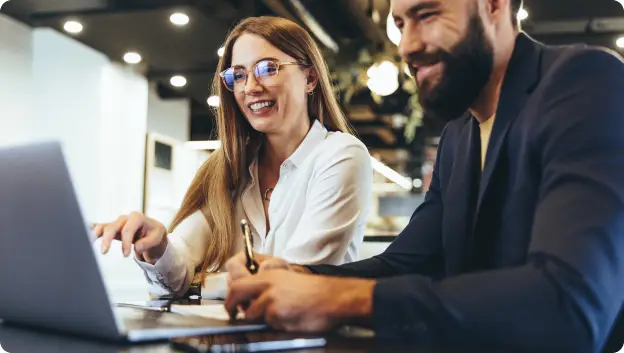 A man and a woman are sitting at a table, both smiling and looking at a laptop computer. The woman, with long hair and glasses, is pointing at the screen, while the man, who has a beard, is holding a pen in his hand. The setting appears to be a modern workspace with warm lighting.