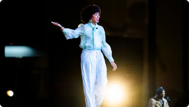 A speaker with curly hair stands on stage in a light blue shirt and white pants, gesturing with her hand while engaging the audience. A figure can be seen on the side, possibly another participant or performer, while bright stage lights illuminate the scene.