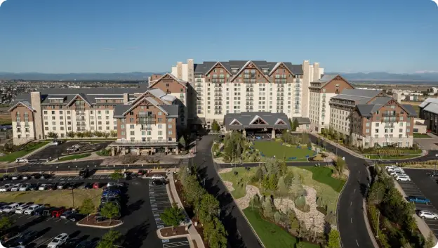 An aerial view of a large hotel complex with a distinctive design featuring multiple peaks on the roof. The foreground includes landscaped gardens and a parking lot filled with vehicles. In the background, there are mountain ranges visible under a clear blue sky. The hotel has stone and wood architectural elements, giving it a rustic yet modern appearance.