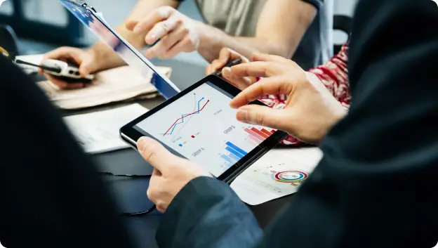 A close-up view of a group working together around a table. A person's hands are holding a tablet displaying various graphs and data points, while other hands reach toward the tablet, indicating collaboration. Papers with charts and notes can be seen on the table, highlighting a busy and engaged workspace atmosphere.
