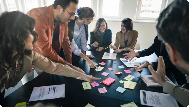 A group of diverse individuals is gathered around a large black table, actively engaging with various sticky notes and papers. Some are pointing at the notes while others are discussing ideas. The atmosphere appears collaborative and dynamic, with bright natural light illuminating the space from the windows behind them.
