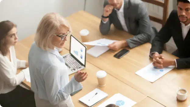 A woman with blonde hair and glasses stands at a conference table speaking to a group of four attendees, who are seated around her. Various documents with charts and graphs are placed on the table, alongside coffee cups. The setting appears to be a professional meeting or presentation environment.