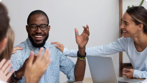 A joyful man wearing glasses is sitting at a table, smiling broadly as he raises his hands in celebration. He is surrounded by two people reaching out to him in a supportive manner, with one person on his left gently touching his shoulder. A laptop is visible on the table in front of them, indicating a collaborative or celebratory moment. The overall atmosphere conveys a sense of happiness and camaraderie among colleagues.