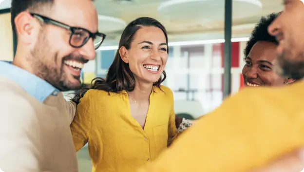 A group of four smiling individuals engages in a lively conversation, expressing joy and camaraderie. They appear to be in a bright, modern workspace, with warm lighting and colorful decor in the background. One woman, wearing a yellow shirt, stands at the center, laughing while two men and another woman lean in, contributing to the friendly atmosphere.