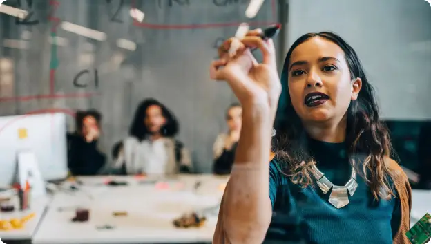 A woman with long hair is standing in a conference room, holding a marker and writing on a glass board. She is wearing a dark shirt and a statement necklace. In the background, several people are seated and watching her speak, indicating an engaged audience. The setting features a modern workspace with a creative atmosphere.