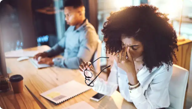 A woman with curly hair is seated at a wooden desk, holding her glasses in one hand while she rubs her temples with the other, displaying a look of stress. In the background, a man sitting at the same table is focused on his laptop. Nearby, there are papers and a coffee cup on the desk. The setting appears to be an office with warm lighting.