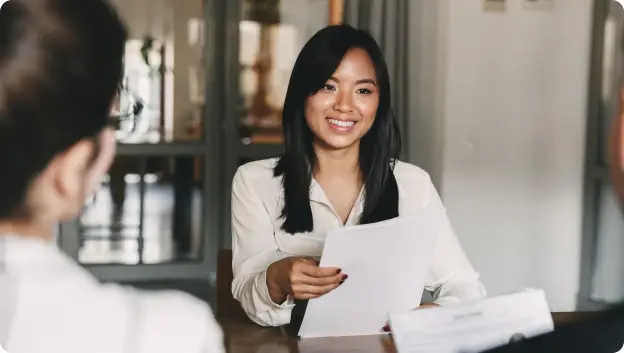A young woman in a white blouse is smiling while seated at a table, holding a piece of paper. She appears to be engaged in conversation with someone out of the frame. The setting is bright and features an office environment in the background.