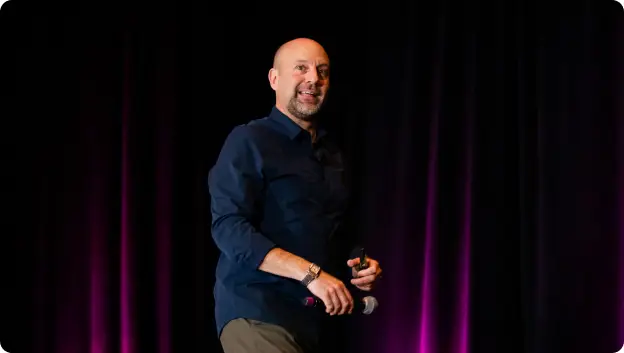 A man smiling and holding a microphone is seen on stage, dressed in a dark blue shirt and light-colored pants. He appears to be engaging with an audience, with a backdrop of soft purple lighting.