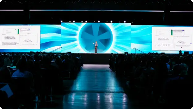 A speaker standing on stage in front of a large audience, presenting against a backdrop featuring vibrant blue graphics and slides with text and visuals. The setting showcases an engaging conference atmosphere, emphasizing interaction and presentation.