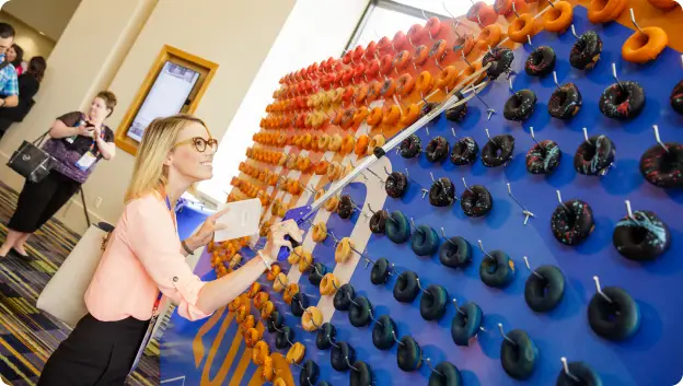 A woman with glasses is using a tool to select a donut from a colorful display wall featuring various types of donuts in multiple colors, including orange, black, and blue. In the background, other conference attendees can be seen engaging in the event.