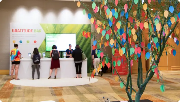 A group of attendees at a Gratitude Bar, with a vibrant tree decorated with colorful leaves in the foreground. The booth features a large green backdrop and individuals interacting at a white counter, likely engaging in activities related to expressing gratitude.