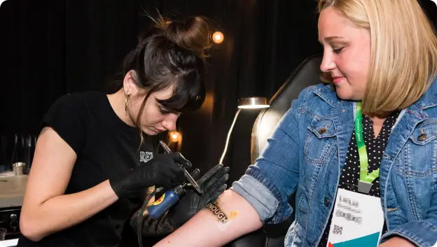 A tattoo artist is carefully applying a tattoo on the forearm of a client, who appears to be enjoying the process. The artist has long dark hair tied back and is wearing black gloves, while the client is wearing a denim jacket and looks pleased. The background features soft lighting and a professional tattoo setup, suggesting a creative and vibrant atmosphere. The word 'ENOUGH' is visible in the tattoo being applied.