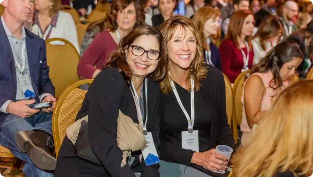 Two smiling women are seated close together at a conference. They are wearing name badges and looking enthusiastically towards the camera. In the background, other attendees are visible, and the setting resembles a conference room with rows of chairs.