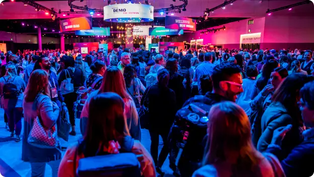 A large crowd gathers at the Workhuman Live conference, with vibrant lighting and various displays in the background. People are engaged in conversation, and large signs can be seen above, including one that says 'DEMO.' The atmosphere is lively, indicating interaction and networking among attendees.