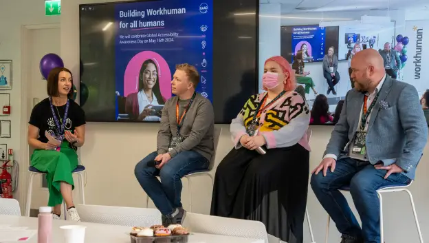 A panel discussion on building an inclusive Workhuman environment, featuring four speakers seated on stools. The background displays a screen highlighting the theme 'Building Workhuman for all humans' along with a photo of a woman smiling, and the event mentions celebrating Global Accessibility Awareness Day on May 18, 2024. The setting includes a table with pastries and balloons, indicating a festive atmosphere.