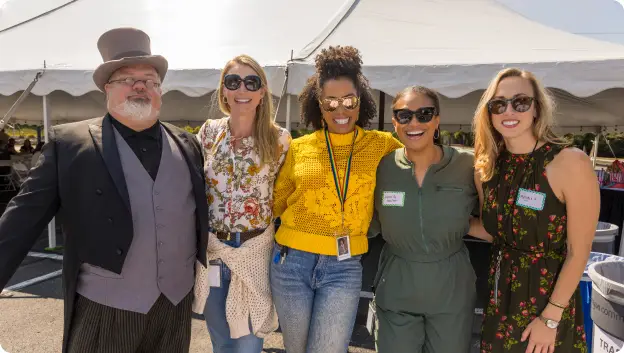 A group of five individuals posing together outdoors at a sunny event under a white tent. The man on the left is wearing a top hat and formal attire, while the other four women are dressed casually in colorful outfits and sunglasses. They appear to be smiling and enjoying the gathering.